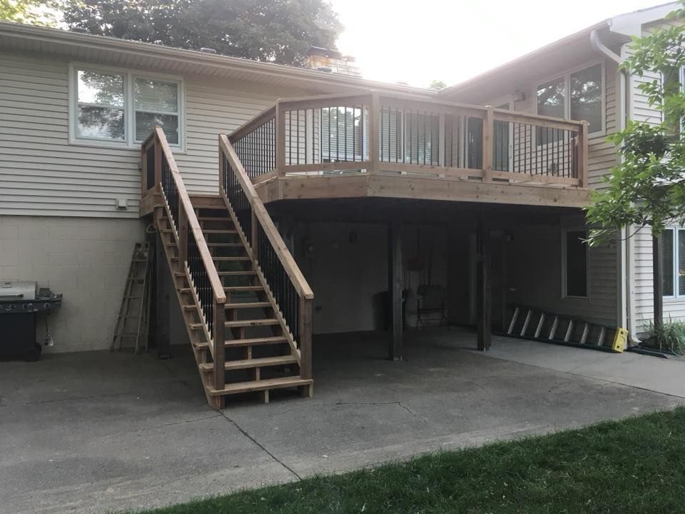 A wooden deck with a staircase leads down to a concrete patio behind a house with tan siding.