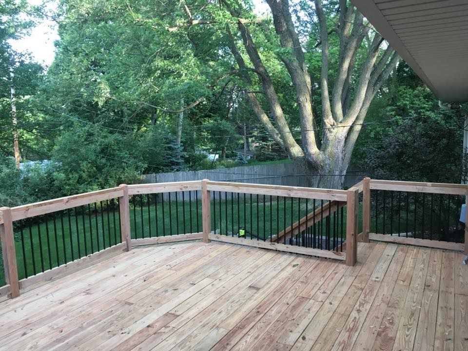 A newly built wooden deck with black metal railings overlooks a lush, tree-filled backyard.