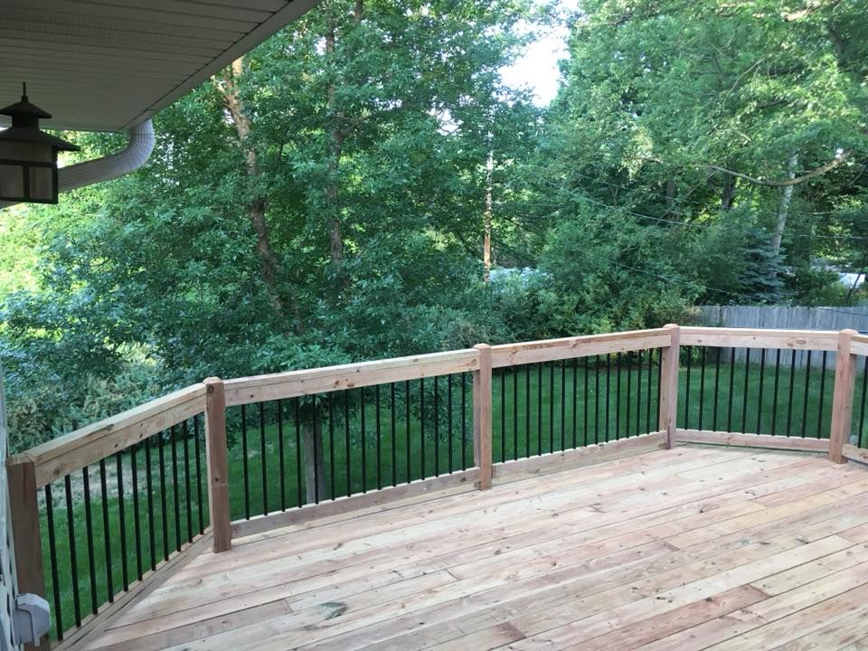 A wooden deck with black metal railings overlooking a lush green yard with trees under a blue sky.