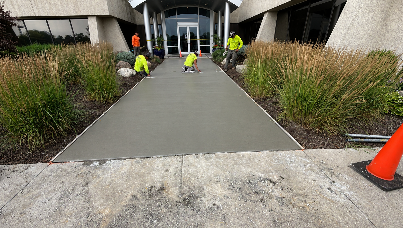 Workers paving a new concrete walkway outside a building entrance, with orange safety cones nearby