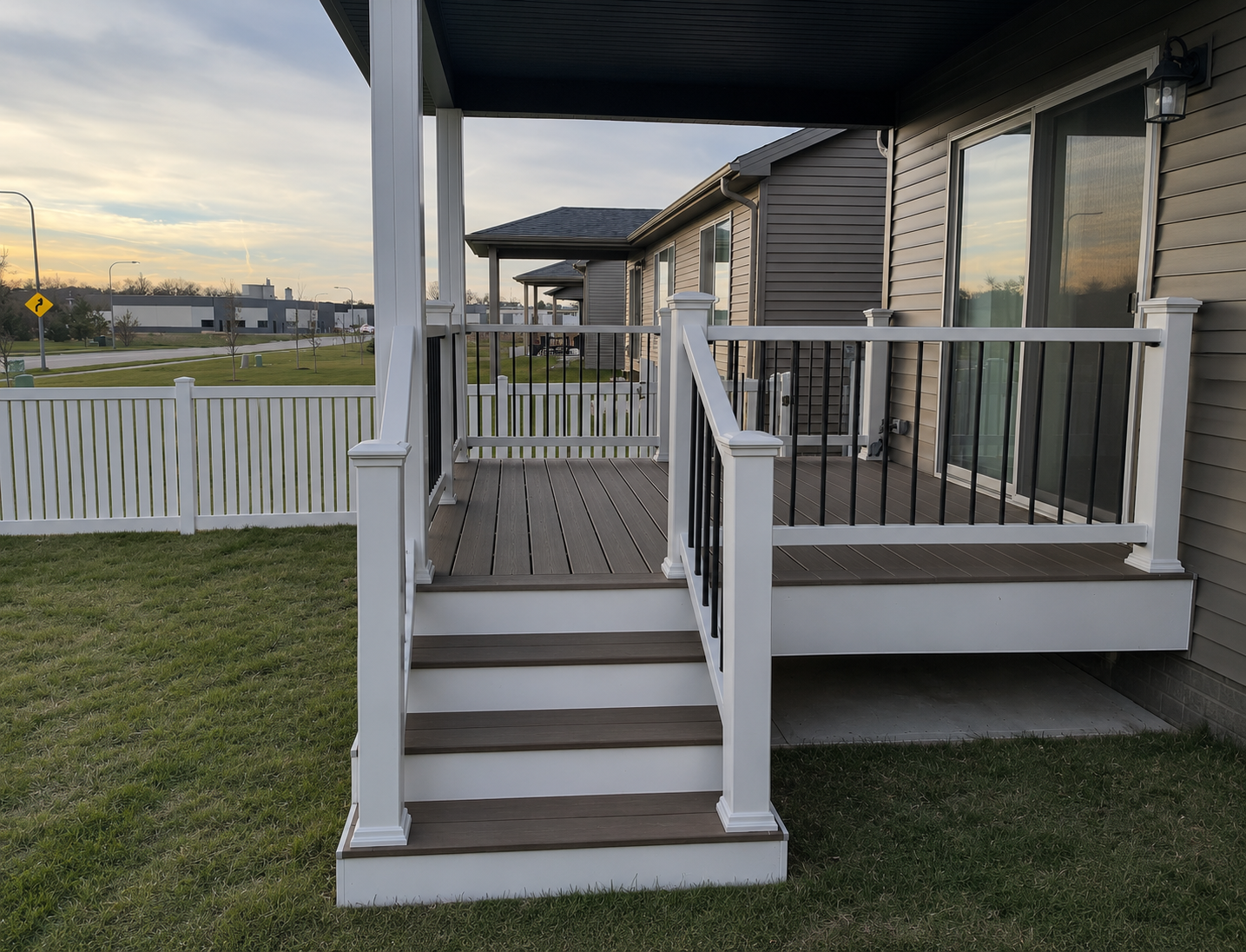 Small front porch with stairs, white railing, and dark siding beside a fenced grassy yard at dusk