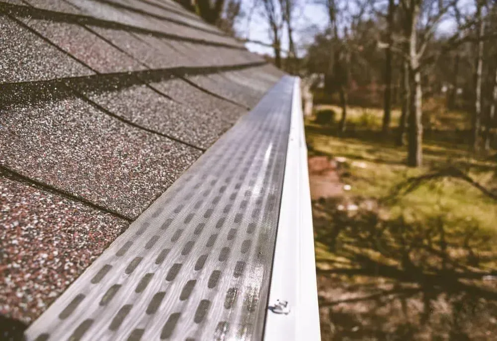 A metal gutter guard with longitudinal slots installed on a residential roof edge, with trees visible in the background.