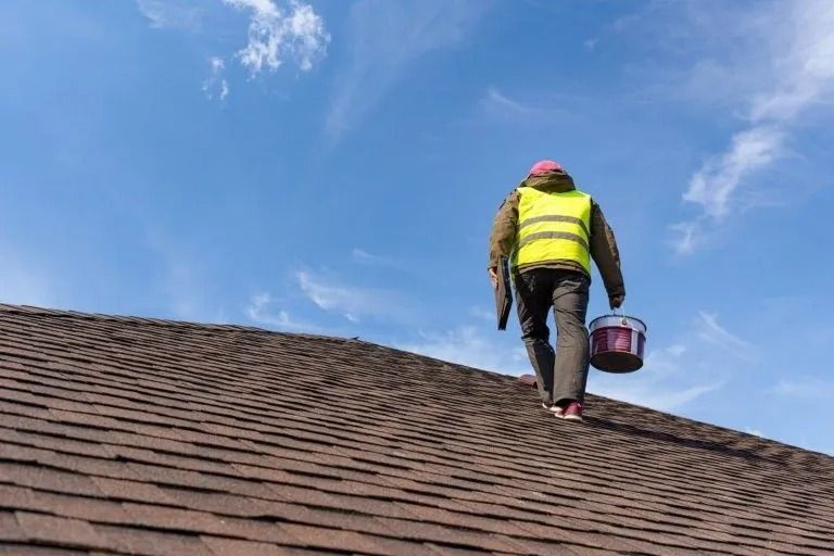 A person wearing a high-visibility vest walks up a brown shingled roof while carrying a bucket against a blue sky.