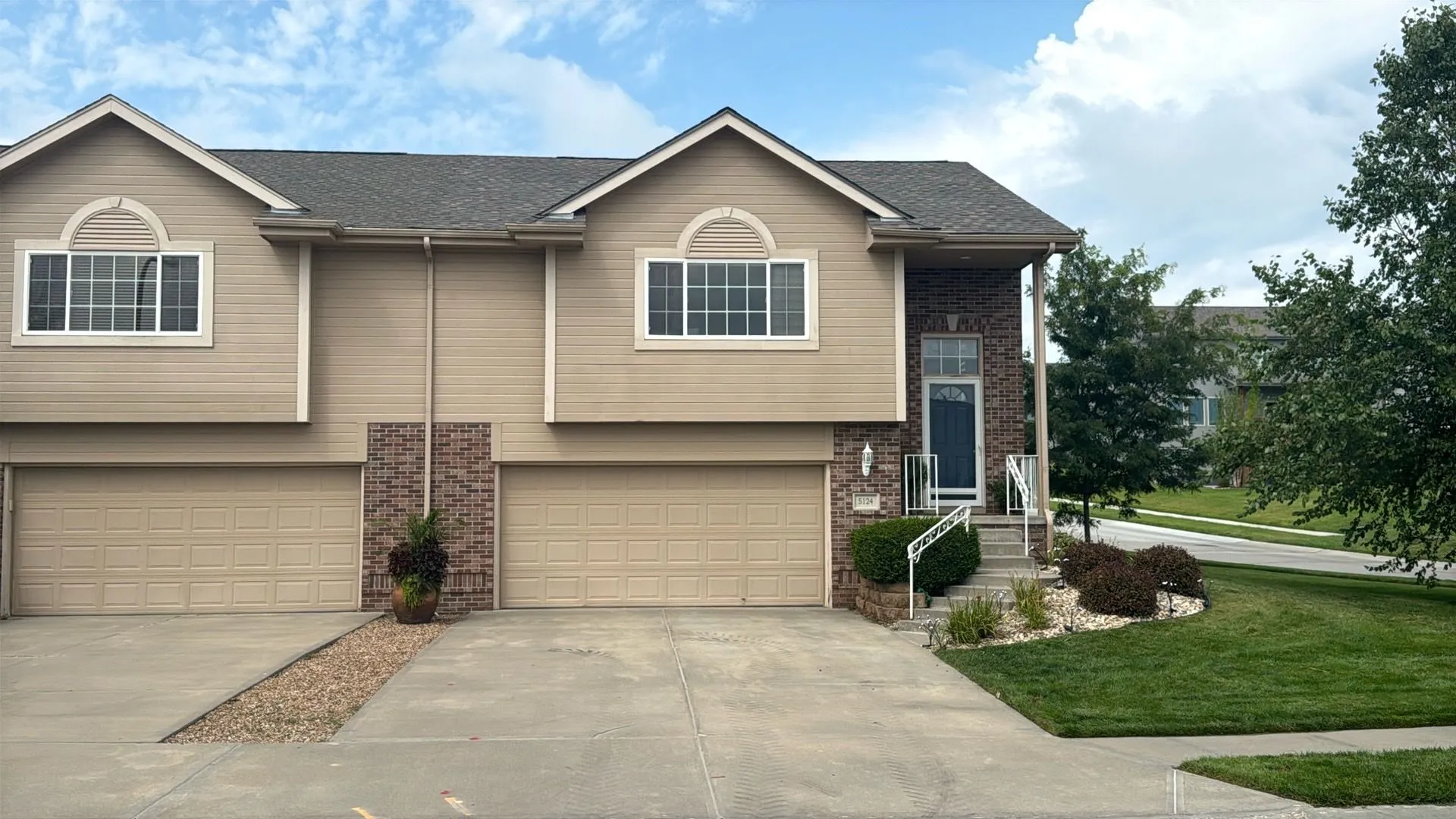 A tan, two-story duplex with two attached garages and a small front garden under a blue, cloudy sky.