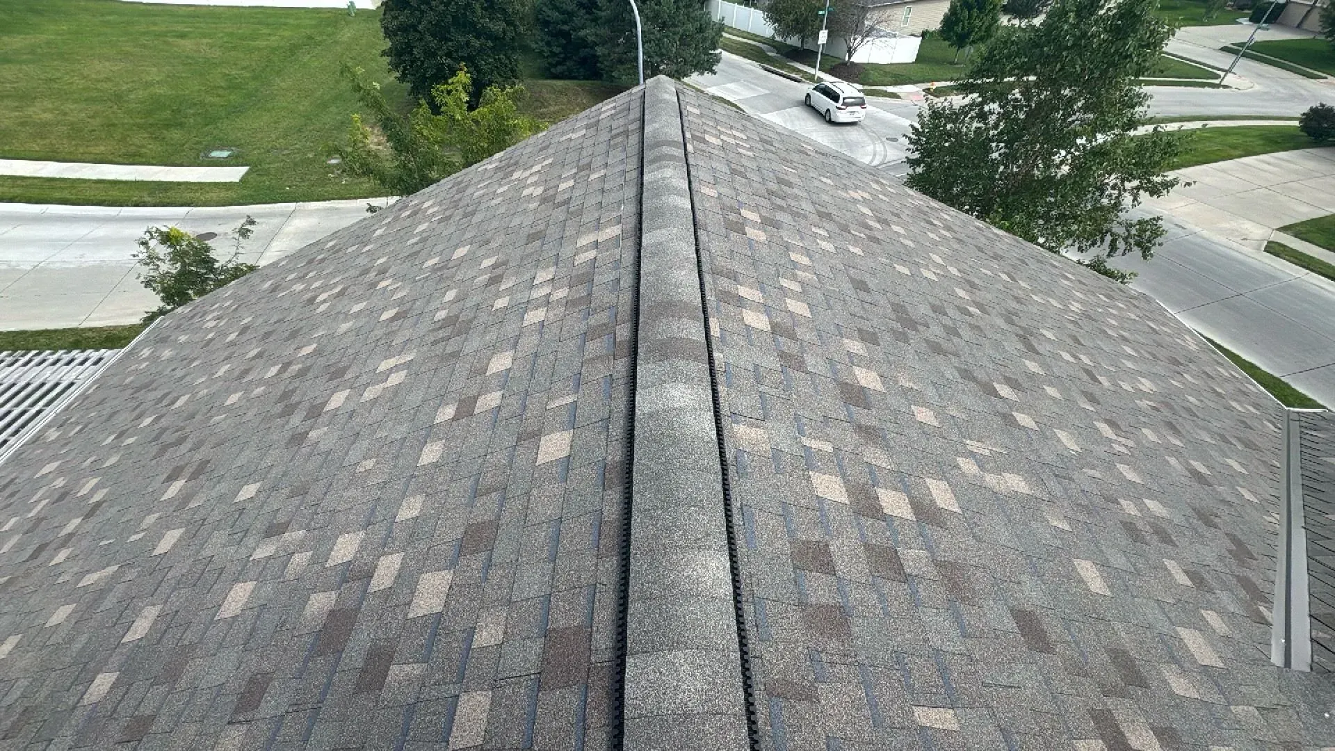 View from a residential roof looking down the shingled ridge line toward a neighborhood street and green lawn.