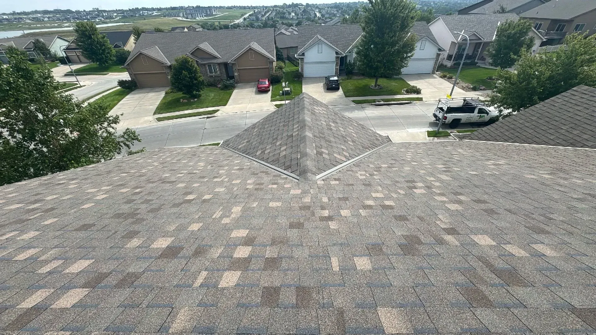 A high-angle view of a grey shingled roof sloping down toward a suburban neighborhood with houses and trees.