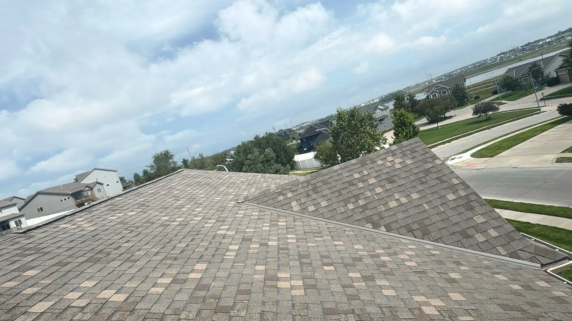 A high-angle view of a grey shingled roof with a peak, overlooking a suburban neighborhood under a cloudy sky.