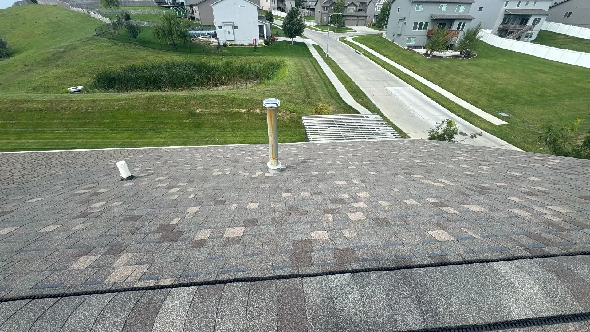 A roof with gray asphalt shingles, two vent pipes, and a suburban street and green landscape in the background.