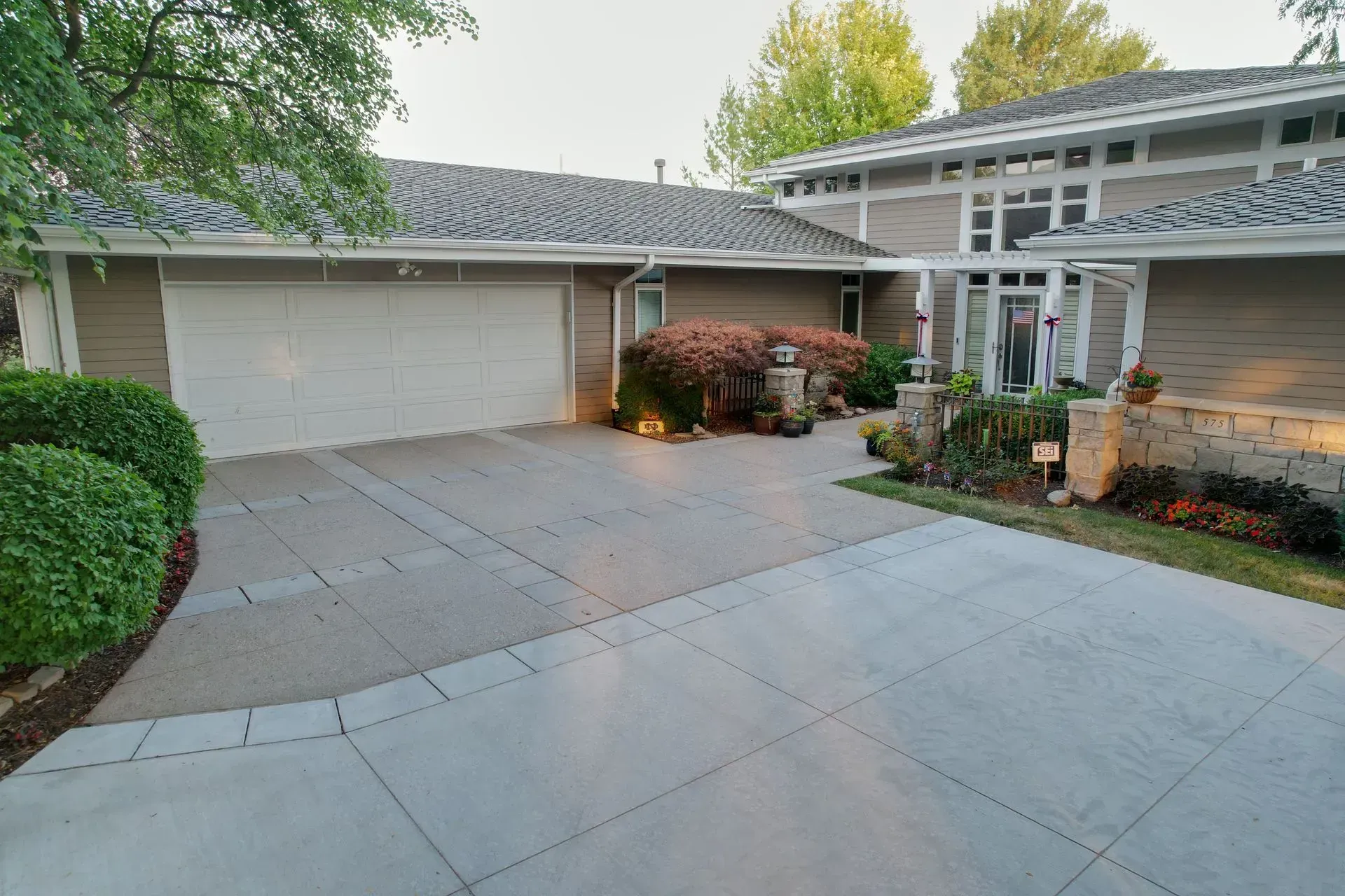 A beige house with a multi-car garage, a gray paved driveway, and landscaping in front of a white framed entryway.