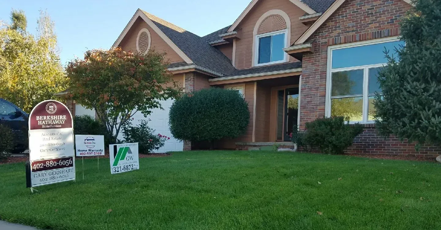 A brick house with a lawn featuring real estate and service signs in a suburban neighborhood.