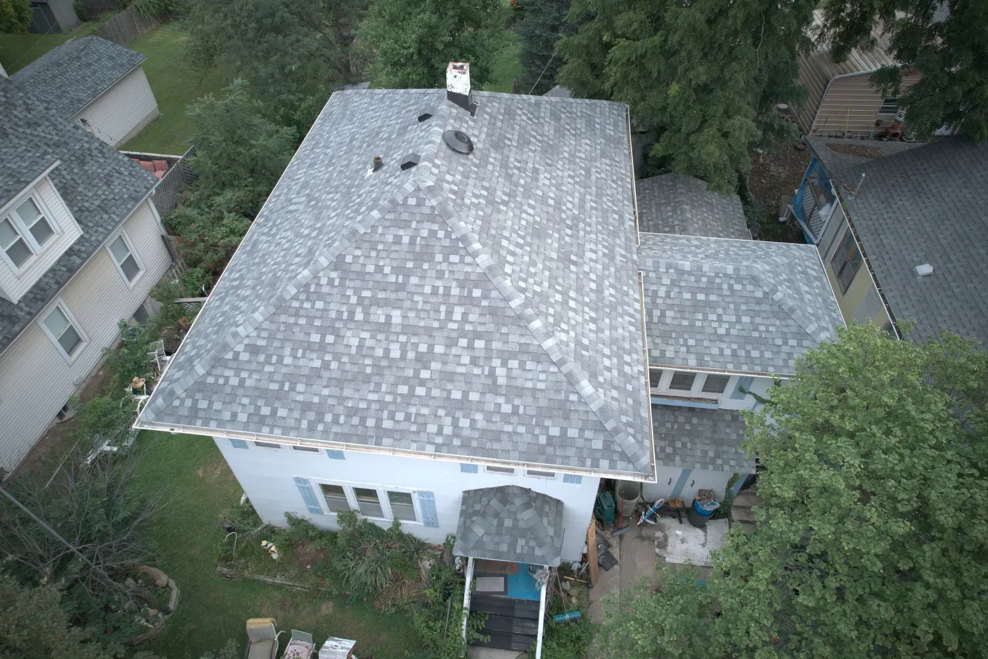 Aerial view of a gray-shingled roof on a white house surrounded by trees in a suburban neighborhood.