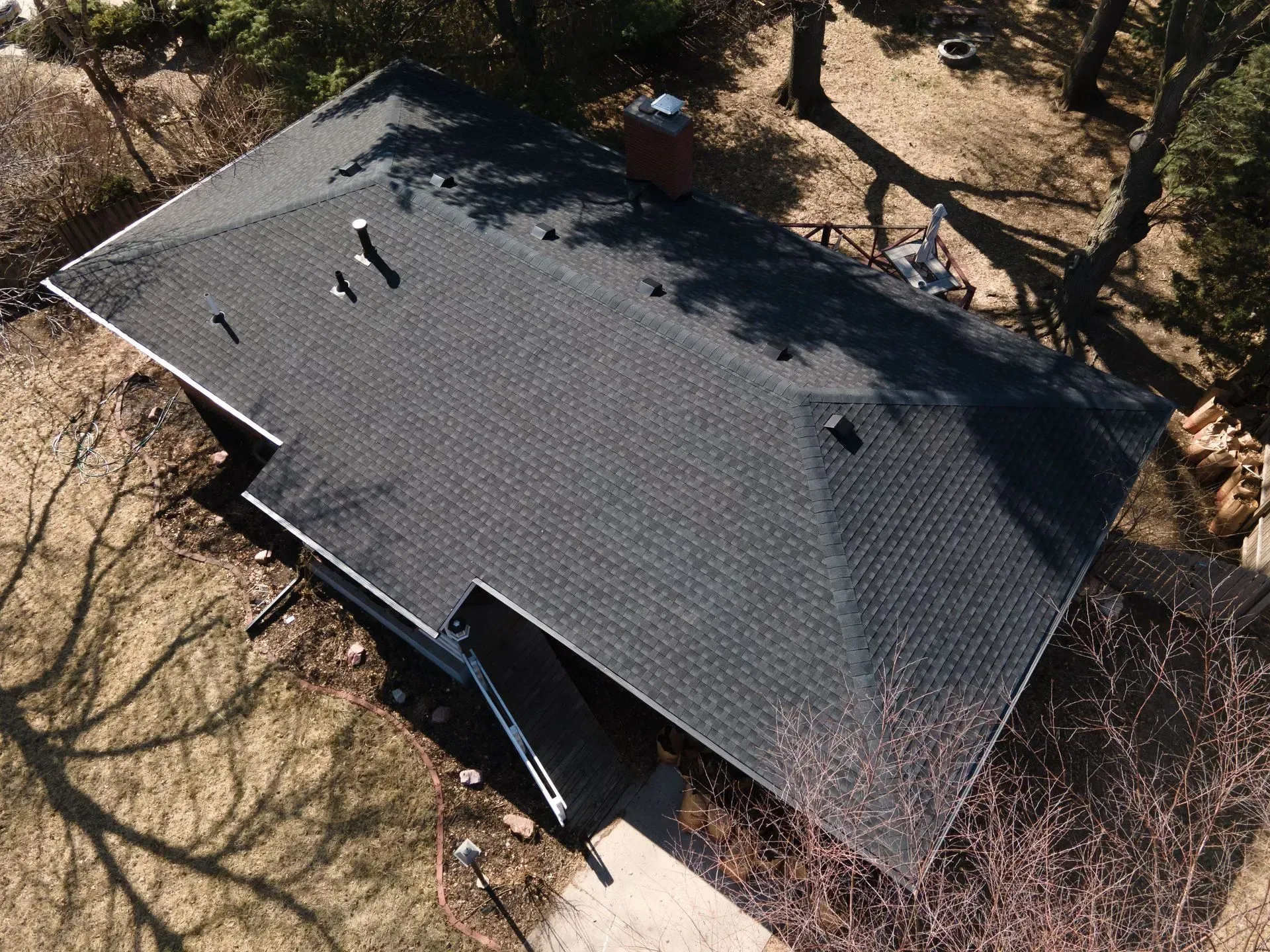 An aerial, high-angle view of a dark-shingled house roof with a chimney, surrounded by a yard with bare trees.
