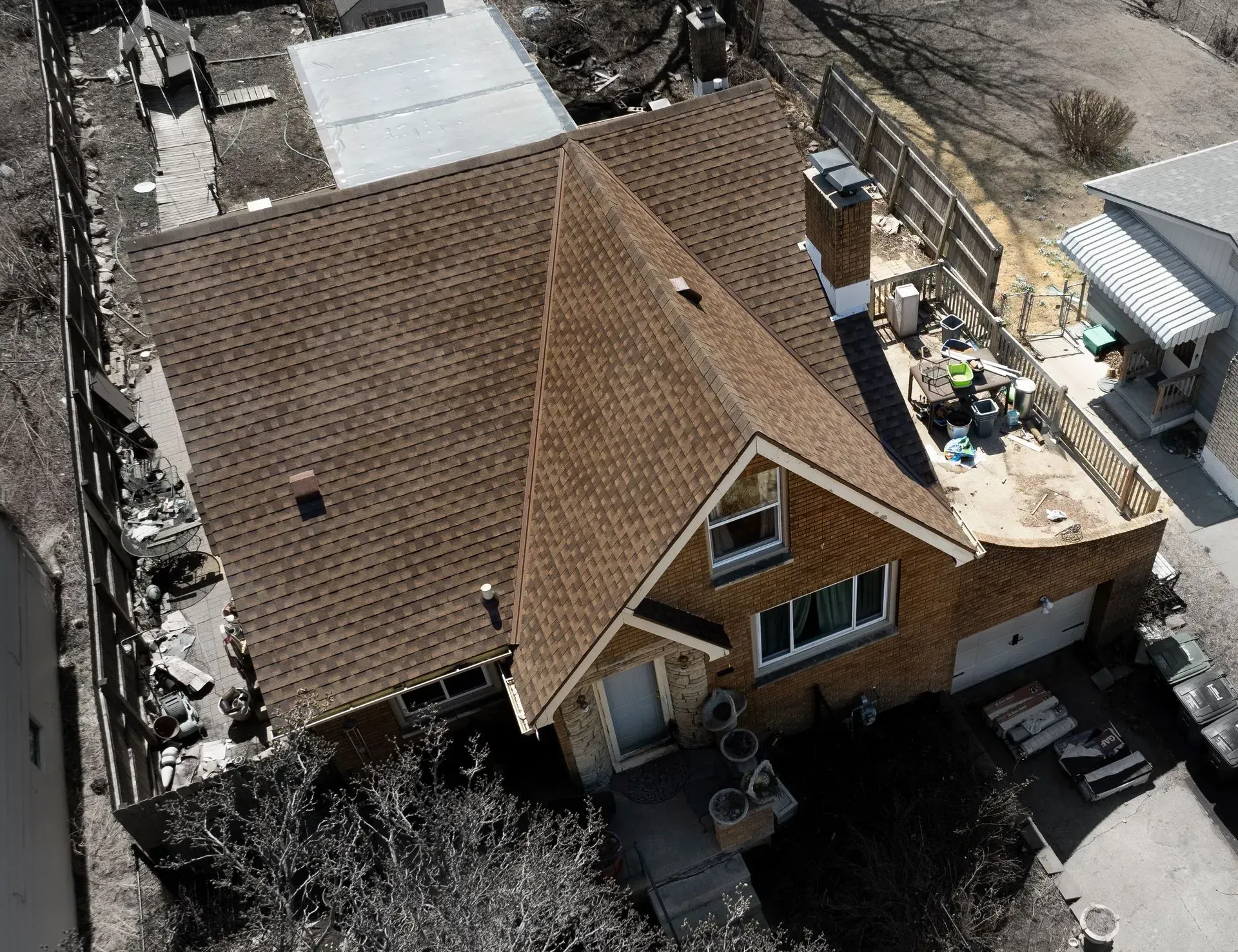 A high-angle aerial view of a single-story brick house with a brown shingled roof, a chimney, and a wooden deck.
