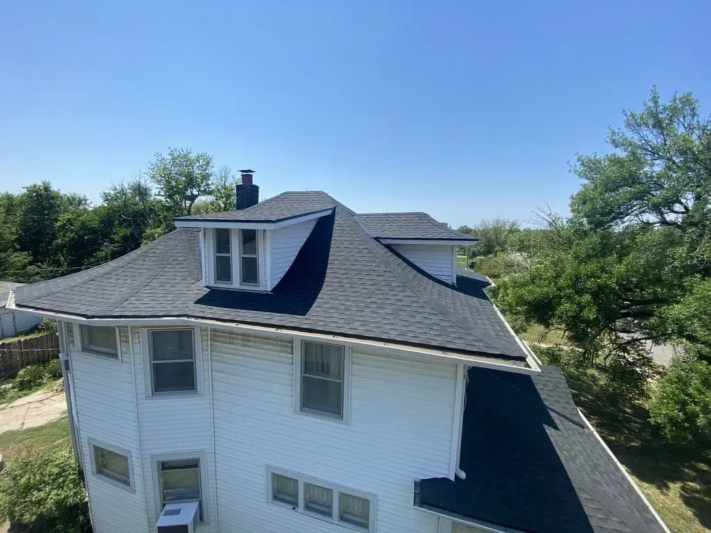 An elevated view of a white, multi-story house with a newly installed dark grey shingled roof under a clear blue sky.