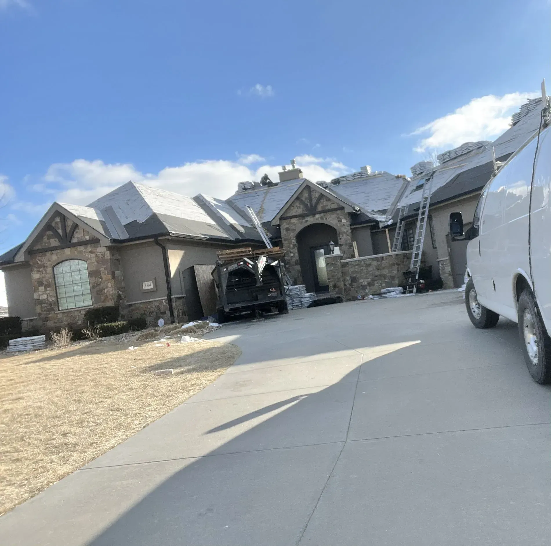 A white van is parked in a driveway in front of a stone-accented house undergoing roof repairs with ladders visible.