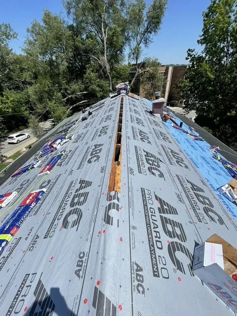 A construction worker installs underlayment on a gabled roof, leaving an open ridge vent gap at the center.