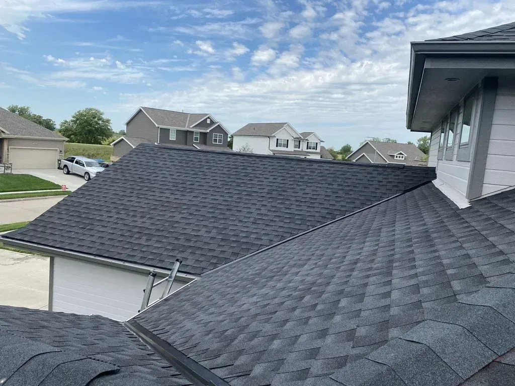 A high-angle view of a newly installed dark grey shingle roof on a suburban house under a partly cloudy sky.