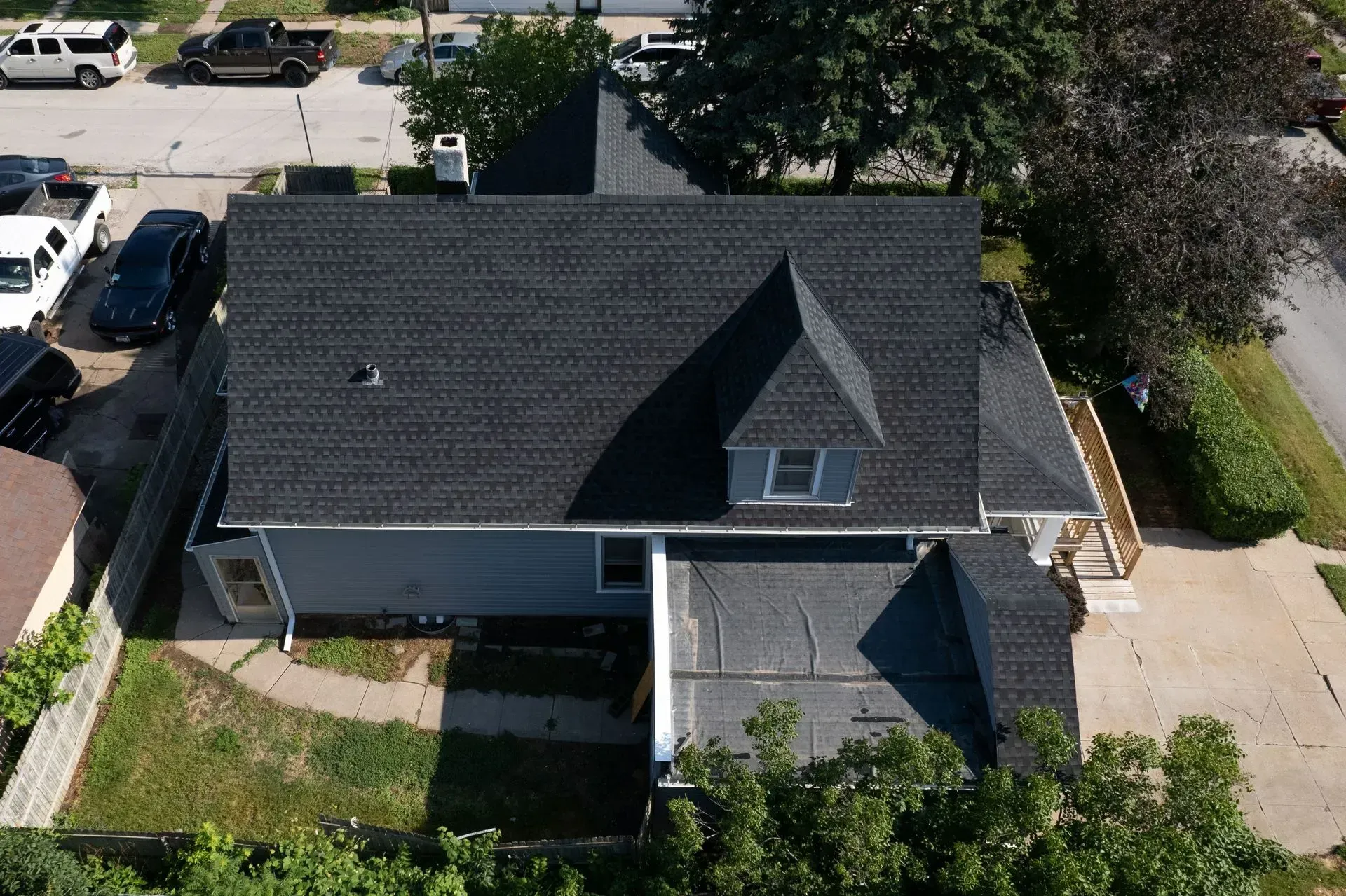 Aerial view of a gray-sided house with a dark shingled roof, a chimney, and a small yard in a residential neighborhood.