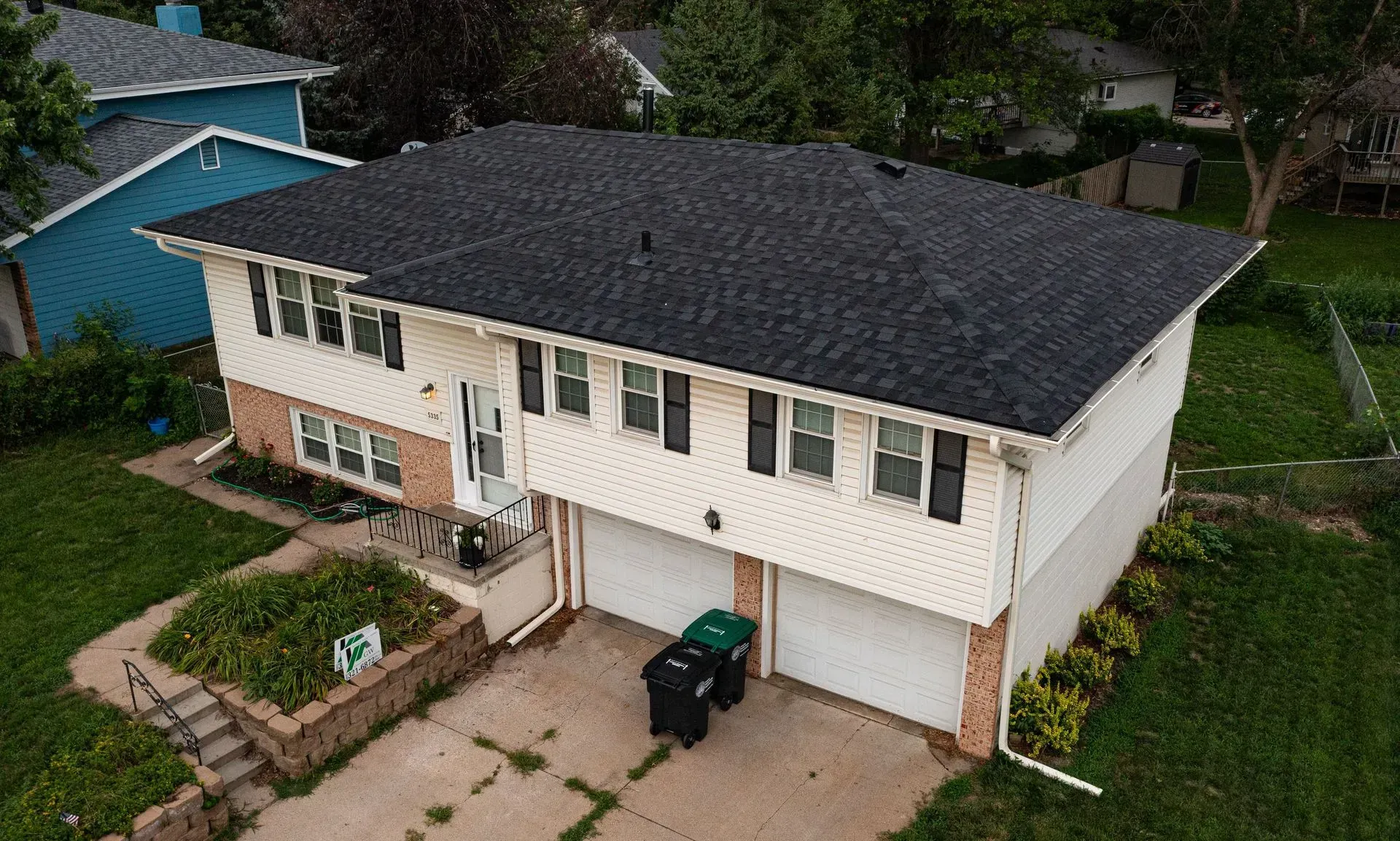 An elevated angle of a split-level home with cream siding, a dark roof, two garage doors, and a front garden.