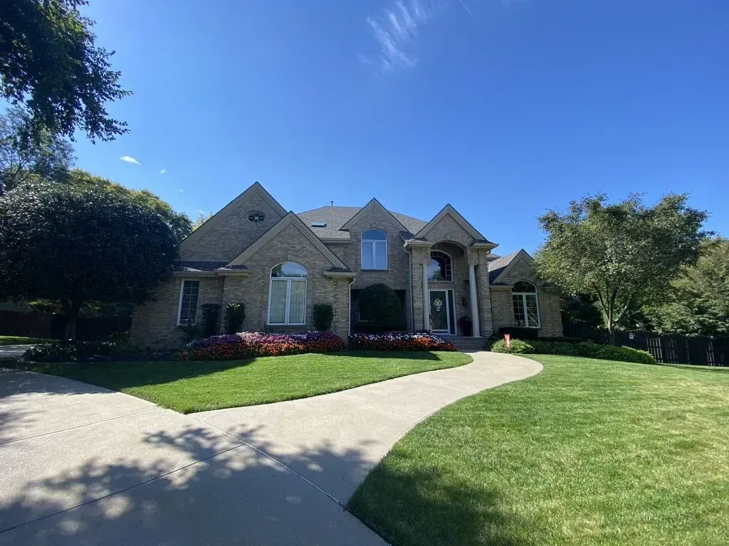 A large, tan brick house with a multi-gabled roof, a front porch with pillars, and a winding driveway on a sunny day.