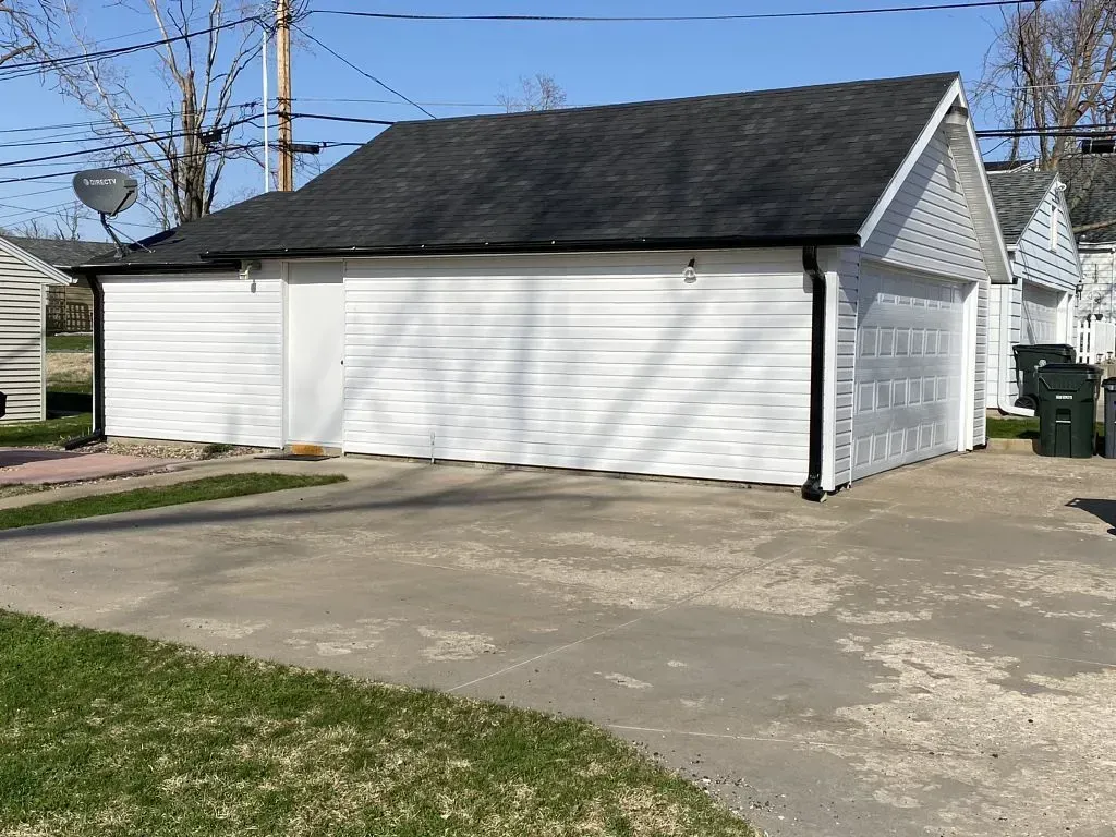 A white detached two-car garage with a dark shingled roof, side entrance door, and a paved concrete driveway.