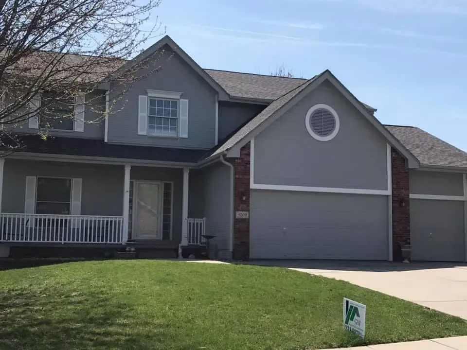 Two-story, gray suburban house with white trim, a front porch, and a multi-car garage under a clear blue sky.