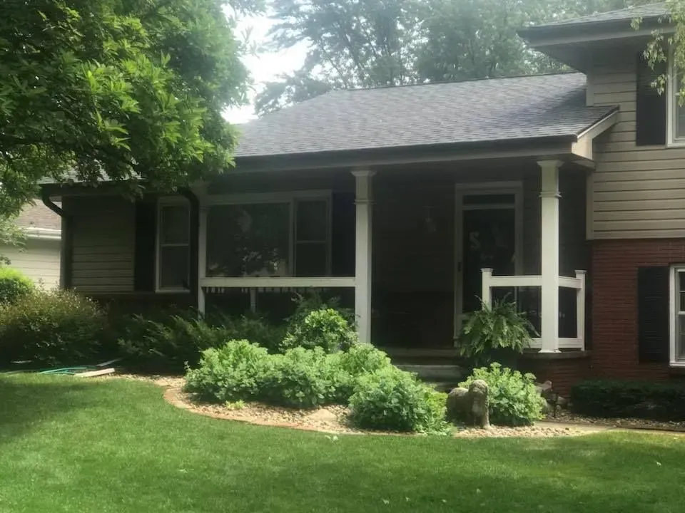 A two-story house with a brick and beige exterior, front porch, and a landscaped front yard with green bushes.