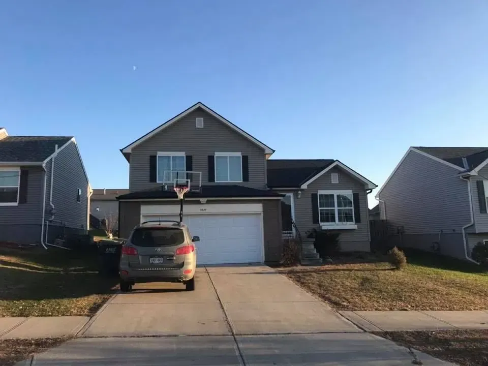 A two-story grey suburban house with a white garage, a basketball hoop, and a silver car parked in the driveway.