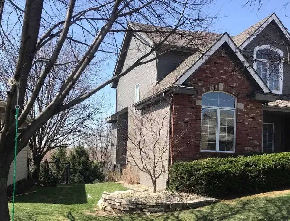 A two-story brick and gray siding house with a stone garden bed and trees in the front yard on a sunny day.