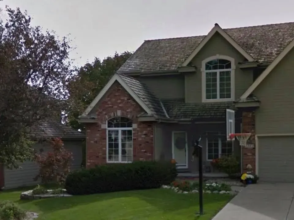 A two-story suburban house with a brick and beige facade, a dark shingled roof, a green lawn, and a driveway.