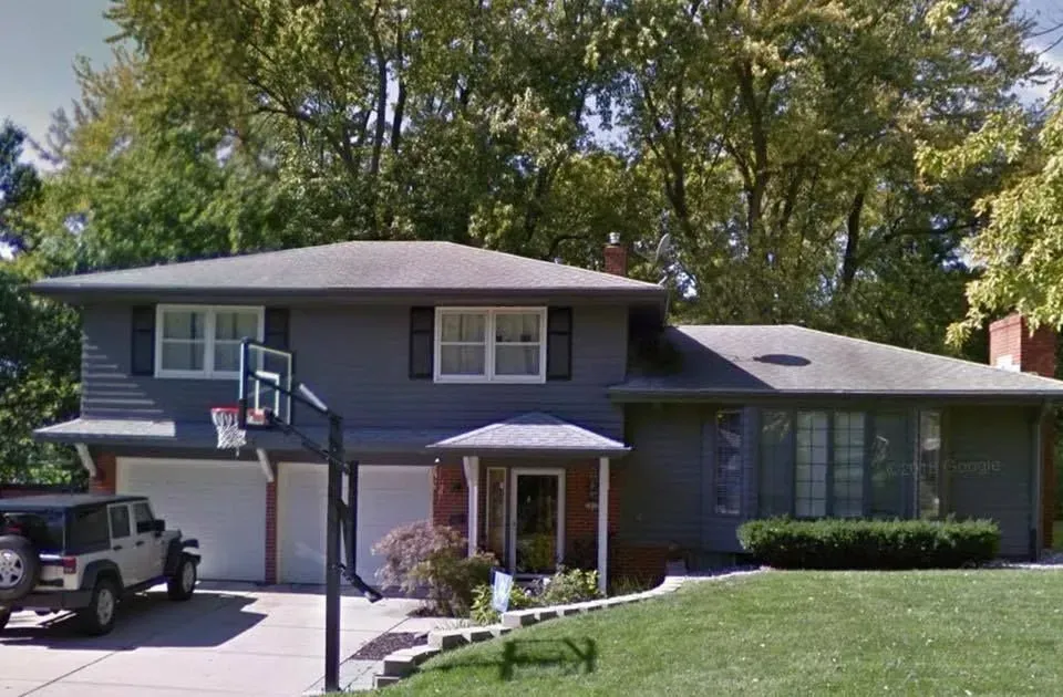 A gray split-level house with a two-car garage, a basketball hoop in the driveway, and a tree-filled front yard.