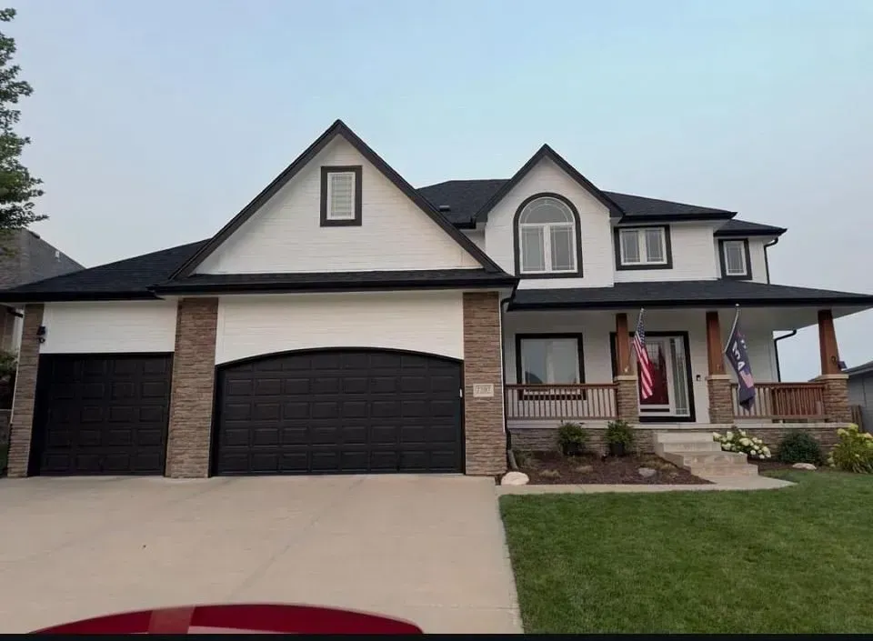 Two-story house with white siding, black roof, arched garage doors, stone accents, and a front porch with American flags.