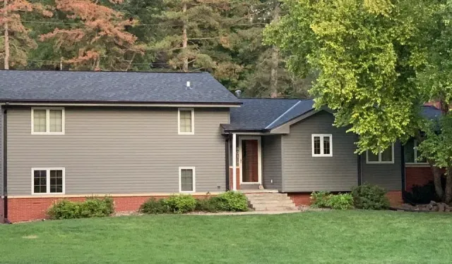 A two-story house with grey horizontal siding, a brick foundation, a dark roof, and a front entry, surrounded by trees.