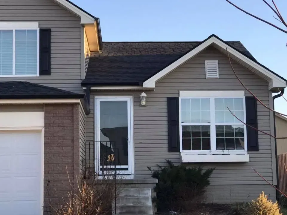 A two-story suburban house with gray horizontal siding, dark roof shingles, a brick accent wall, and black window shutters.
