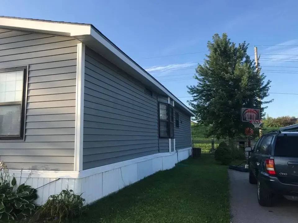 Side view of a grey manufactured home with white skirting, a small window, and a dark SUV parked on a paved driveway.