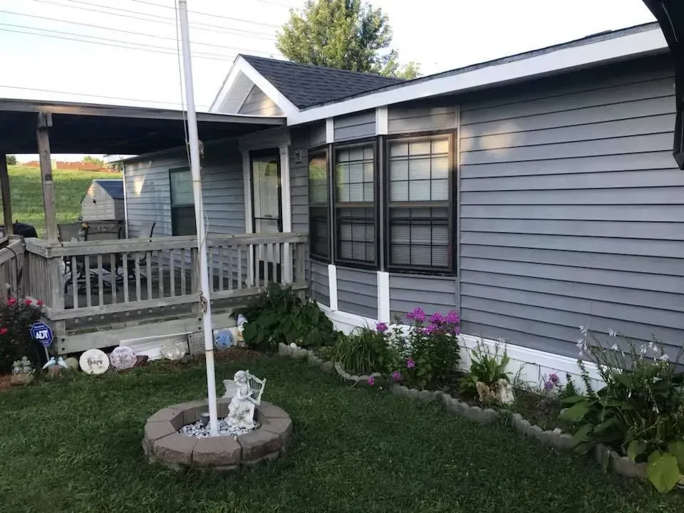A blue-grey mobile home with a front deck, a flag pole in a stone flower bed, and garden landscaping in the yard.