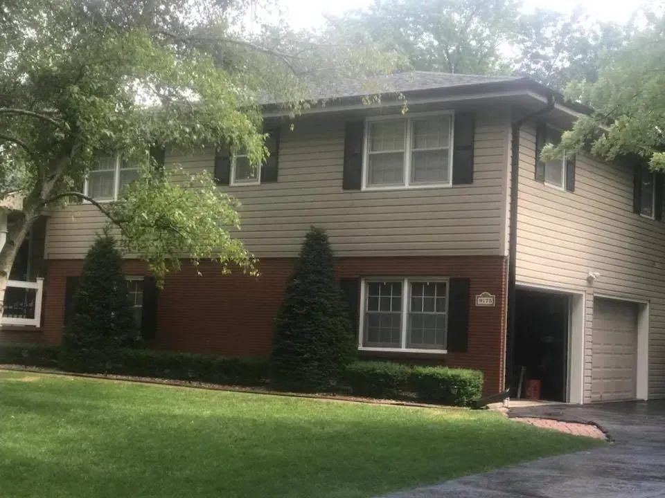 A two-story house with tan siding on the upper level, brown brick on the lower level, and an attached two-car garage.