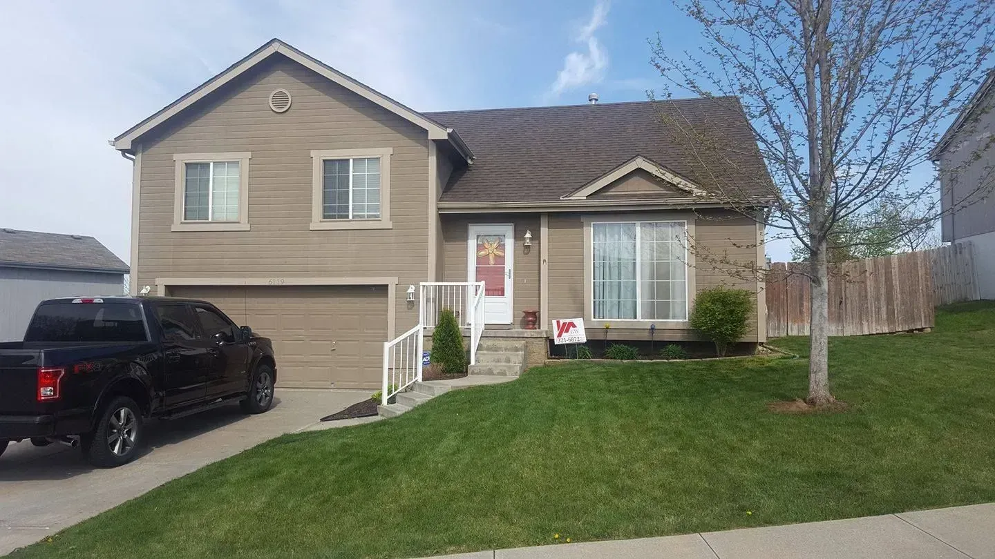 A two-story, tan-sided house with a dark roof and an attached garage, with a black truck parked in the driveway.