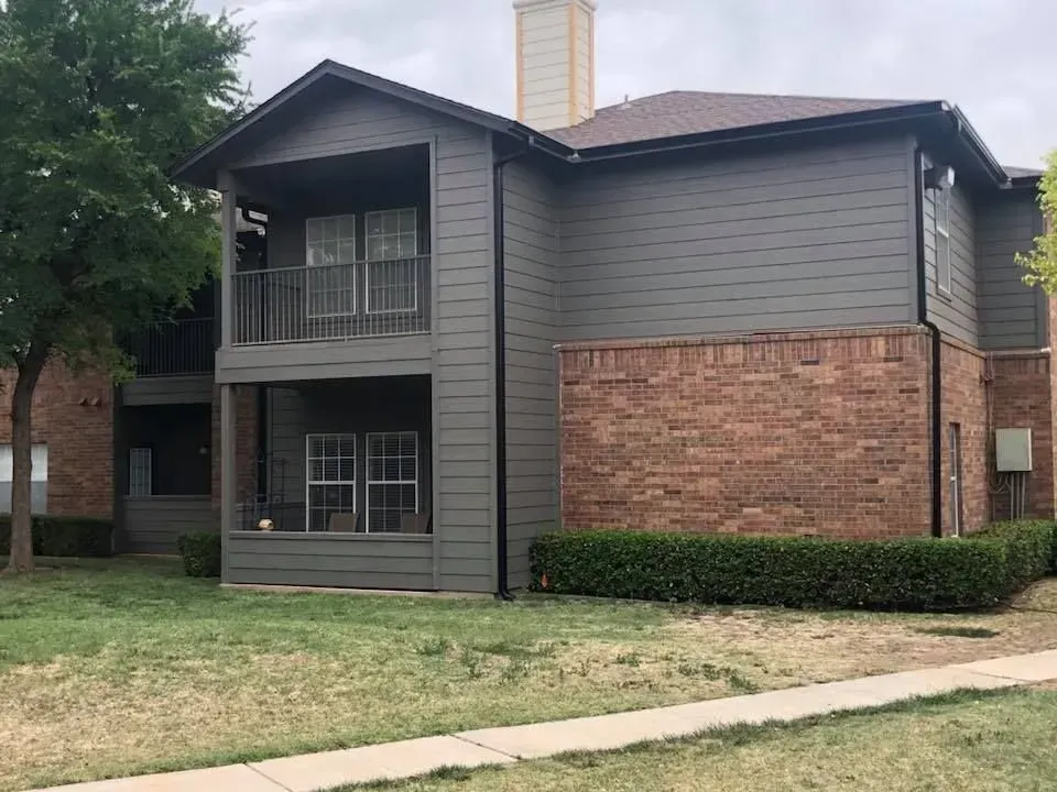 A two-story apartment building with dark gray horizontal siding and a red brick base, surrounded by green landscaping.