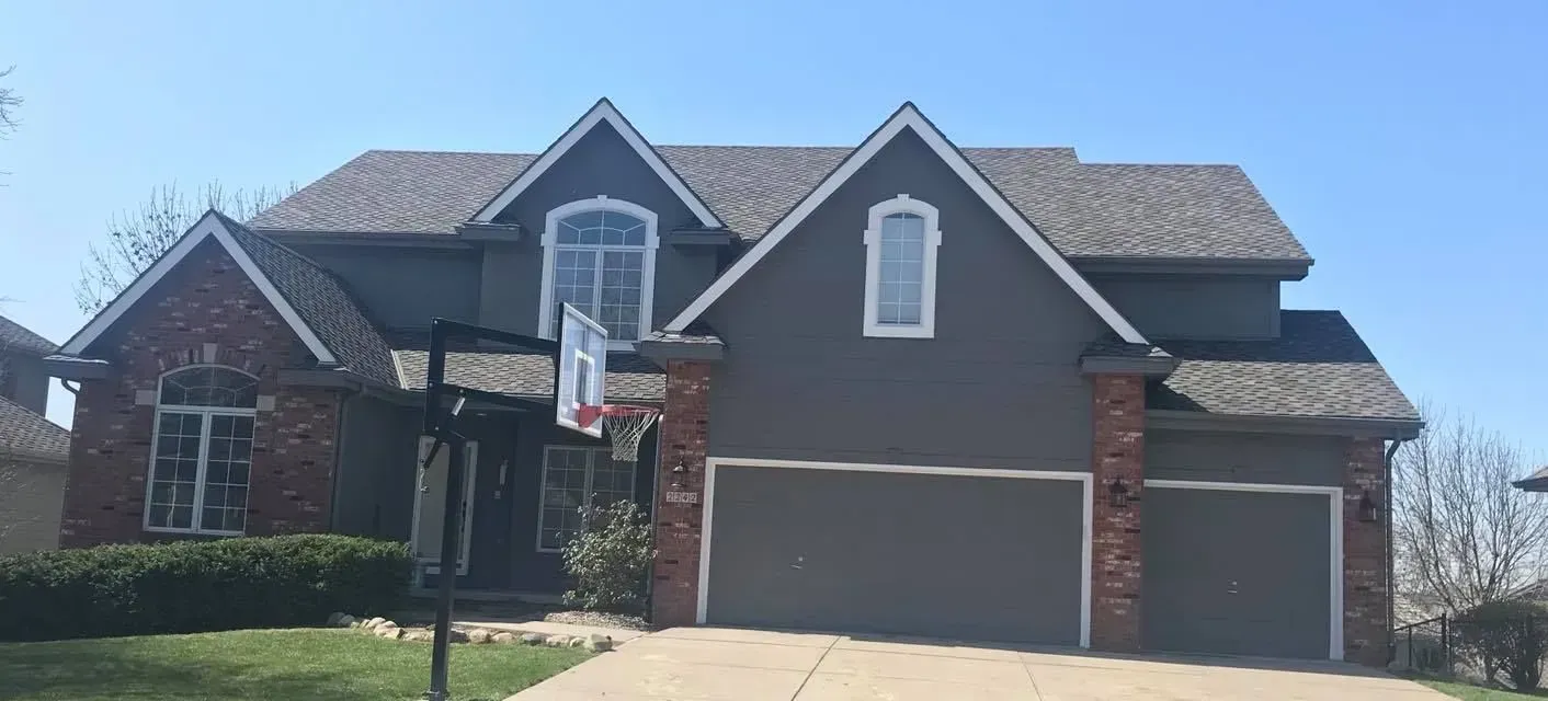 A two-story house with dark gray siding, brick accents, a gabled roof, and a three-car garage under a clear blue sky.