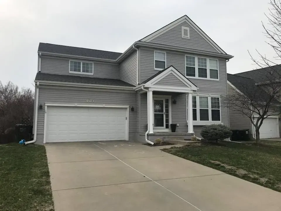 A two-story grey vinyl-sided house with a white garage, front porch, and concrete driveway under an overcast sky.