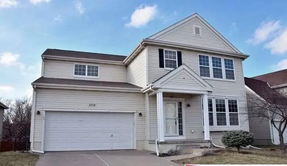 A two-story suburban house with beige siding, a two-car garage, and a covered front porch under a blue sky.