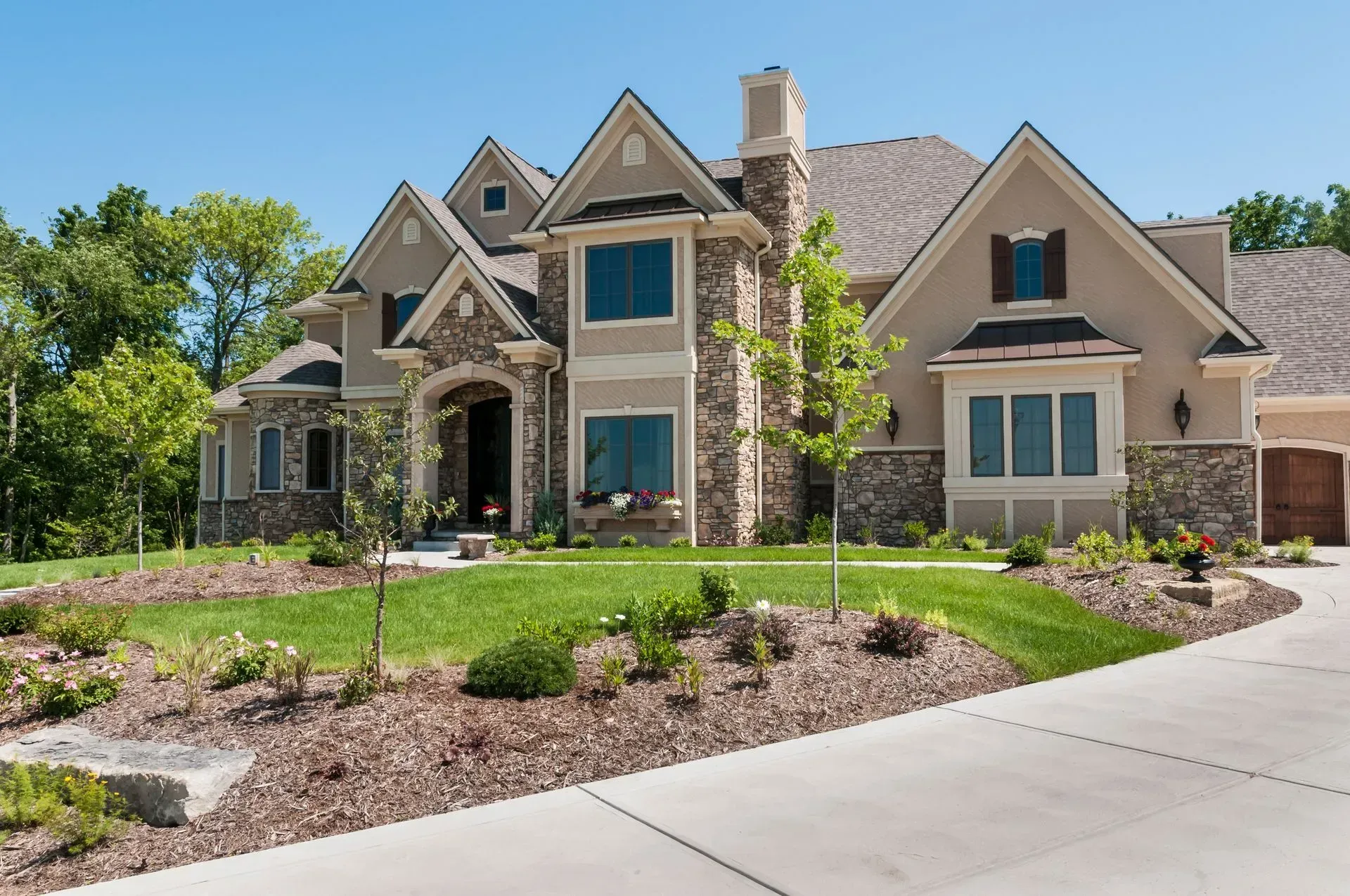 A large, tan suburban house with stone veneer, a steep roof, and a green lawn under a clear blue sky.