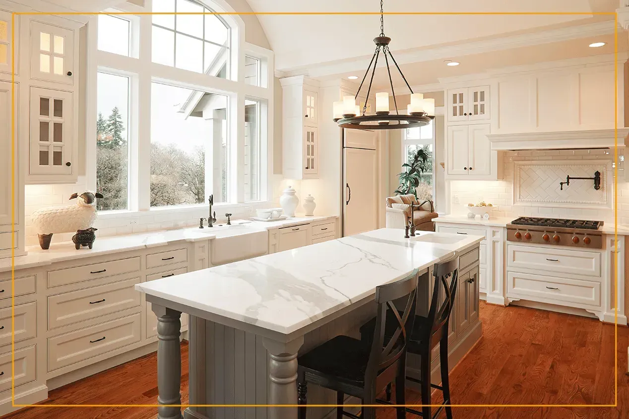 A bright, white kitchen with a large gray island, marble countertops, pendant lighting, and hardwood flooring.