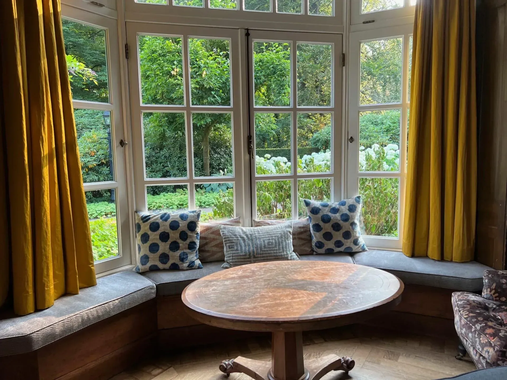 A curved window nook with a cushioned bench, patterned pillows, mustard curtains, and a round wooden table.