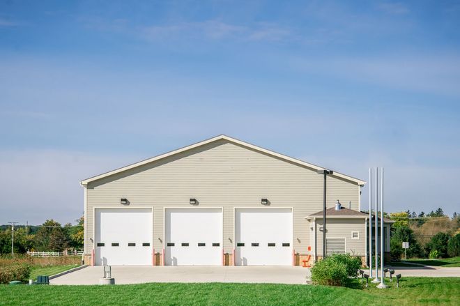 Large beige garage with three white doors and a small side shed on a grassy lot under a blue sky