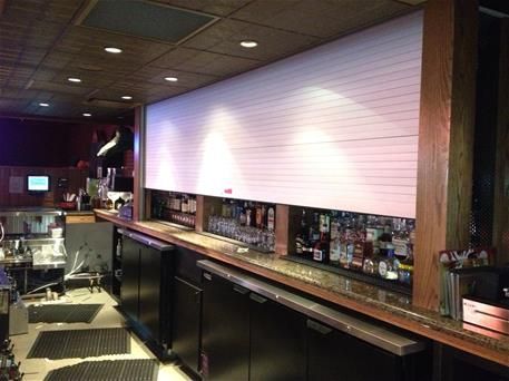 Empty bar interior with a long counter, bottles on shelves, and warm wood paneling