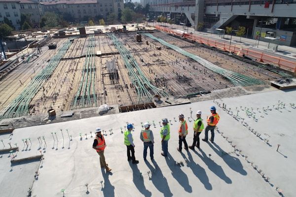 Construction workers in hard hats stand on a concrete slab overlooking a large building site.