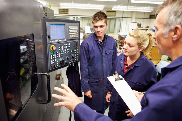 People in blue uniforms inspecting a machine control panel in a factory.