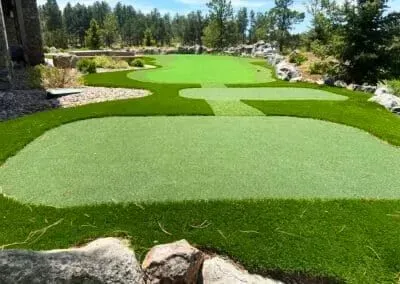 A backyard putting green with synthetic turf, bordered by rocks and a forest landscape under a clear blue sky.